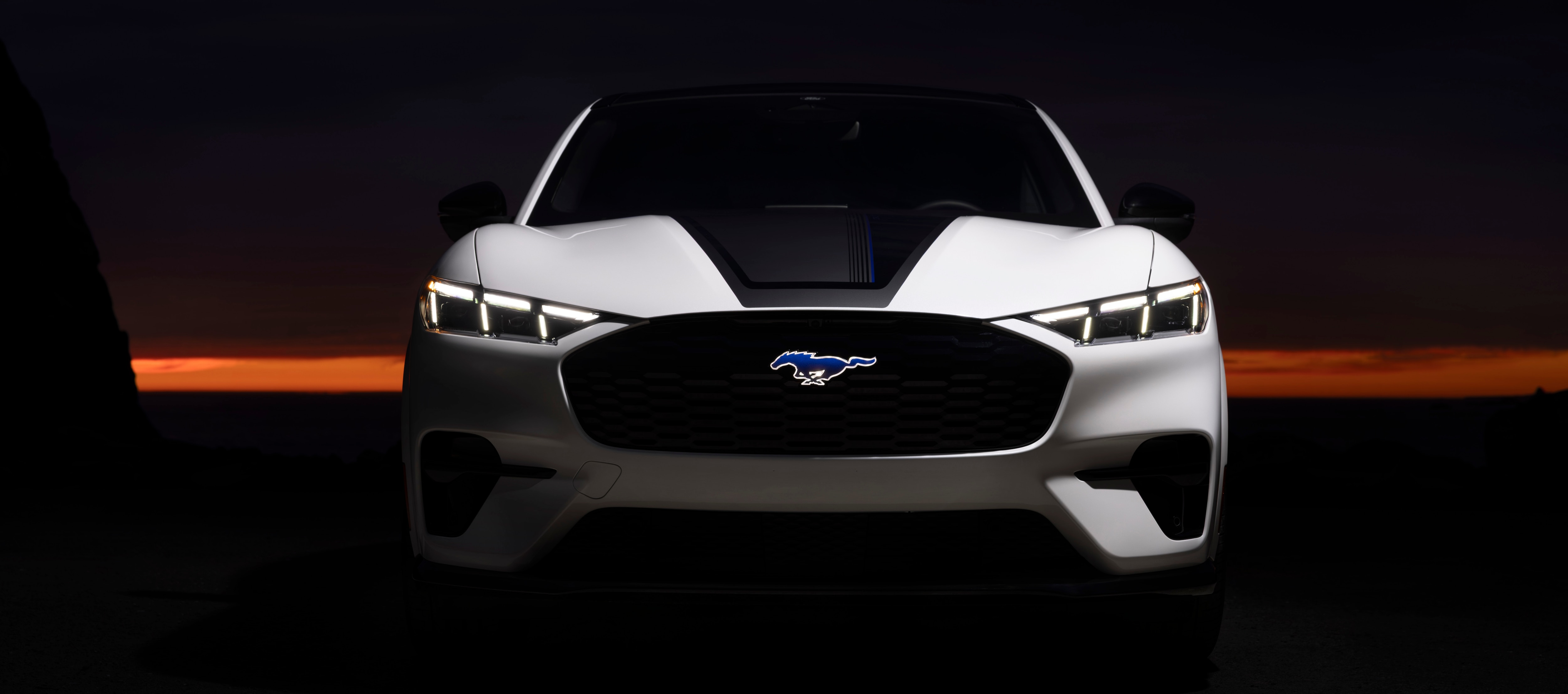 A white Mustang Mach-e is parked in front of a bright orange sunset.